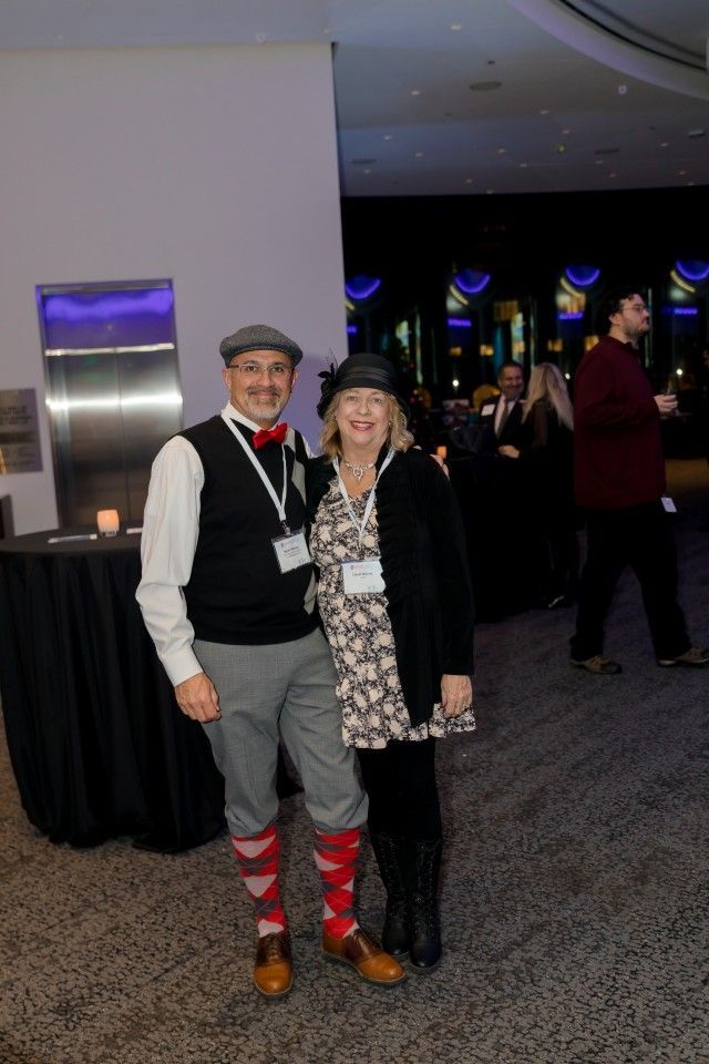 Couple in formal attire, posing at an event. Man in vest, cap, and argyle socks; woman in dress, hat, and cardigan.