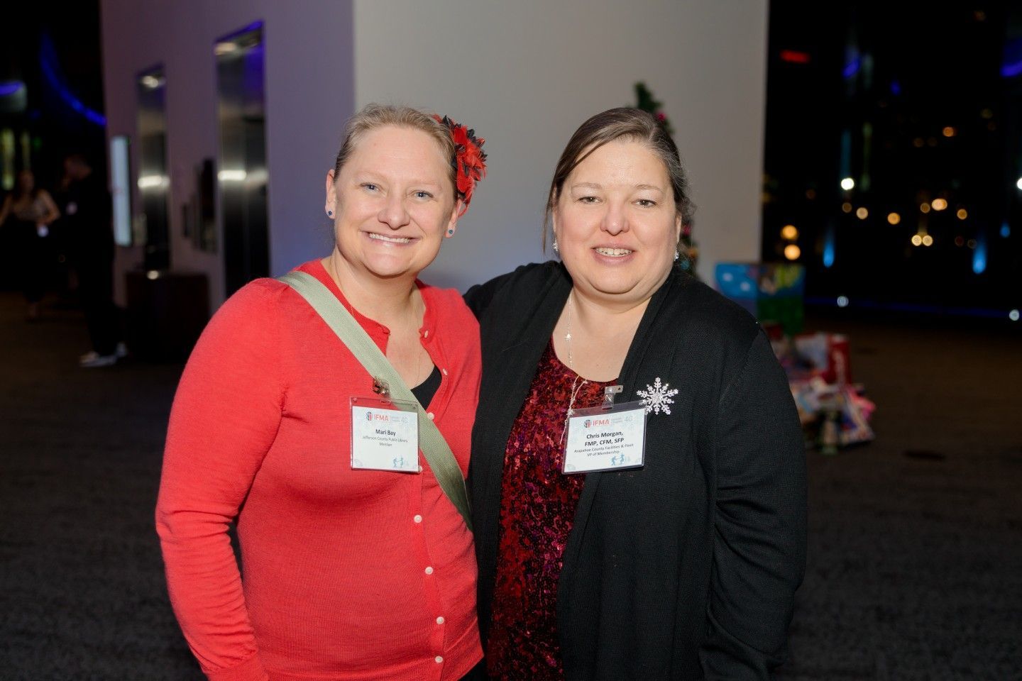 Two women smile, one in red sweater with flower, other in black cardigan over sequin top, at event.