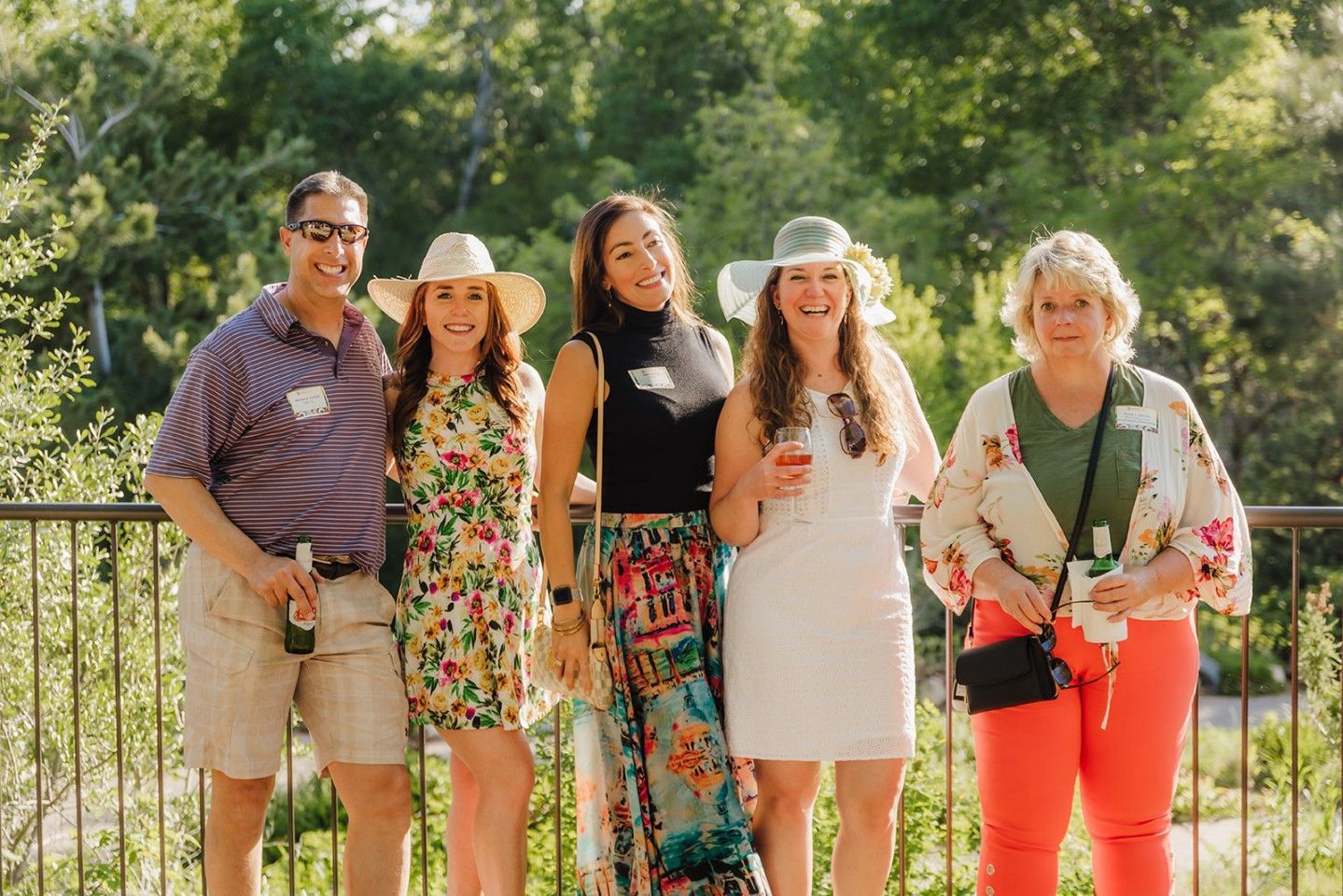 Group of five people smiling outdoors, near a railing, wearing summer attire and hats.