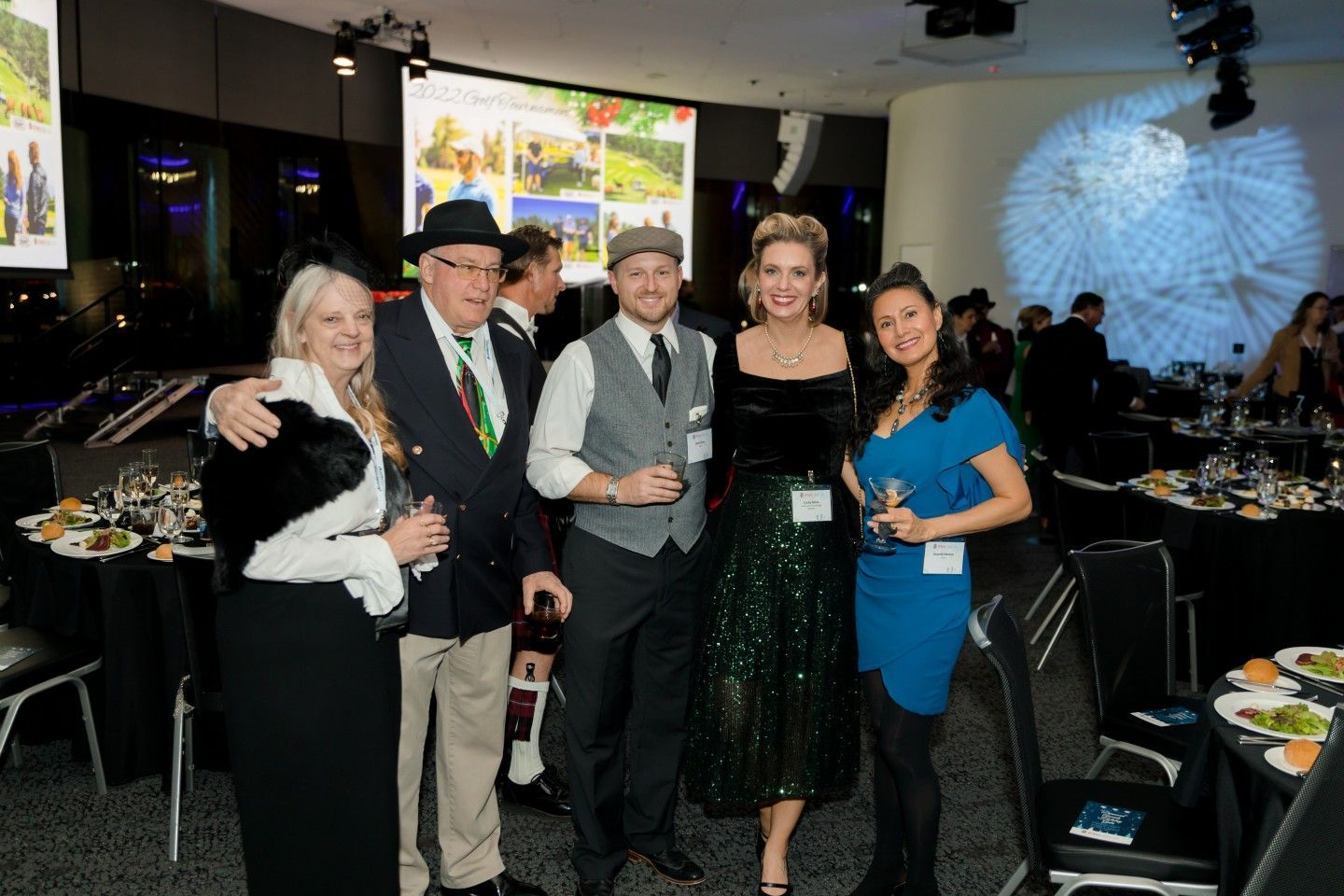 Group of five people at an event, posing with drinks. One man wears a hat and vest.