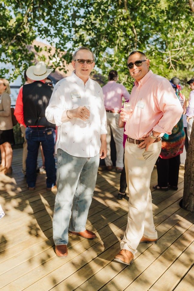 Two men stand on a wooden deck, smiling. One in white shirt, jeans, and sunglasses, other in pink shirt and khakis.