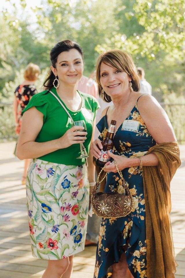 Two women smiling at an outdoor event. One in green top and floral skirt, the other in a floral dress with a shawl.