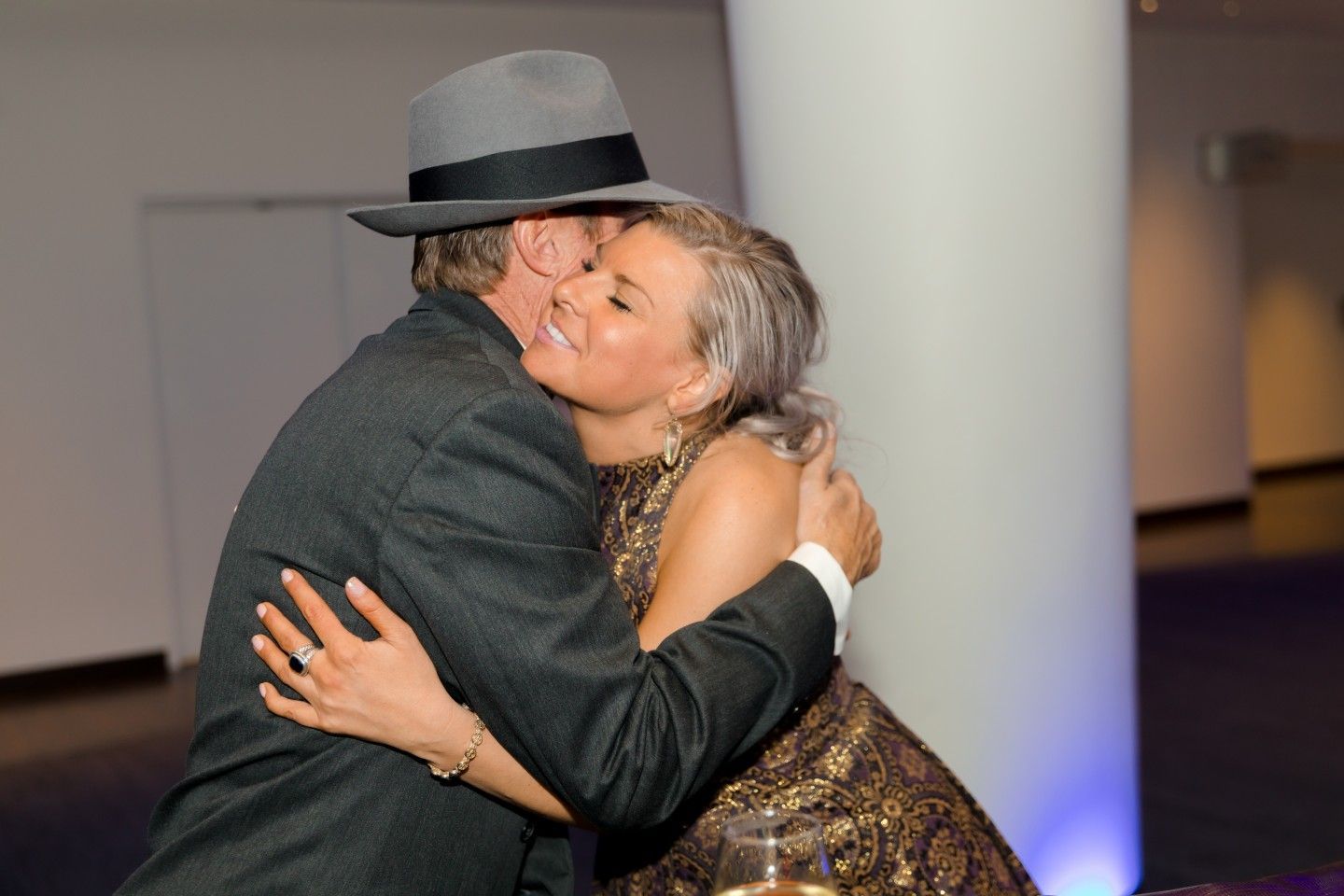 Man in fedora embraces woman in gold dress; interior setting.