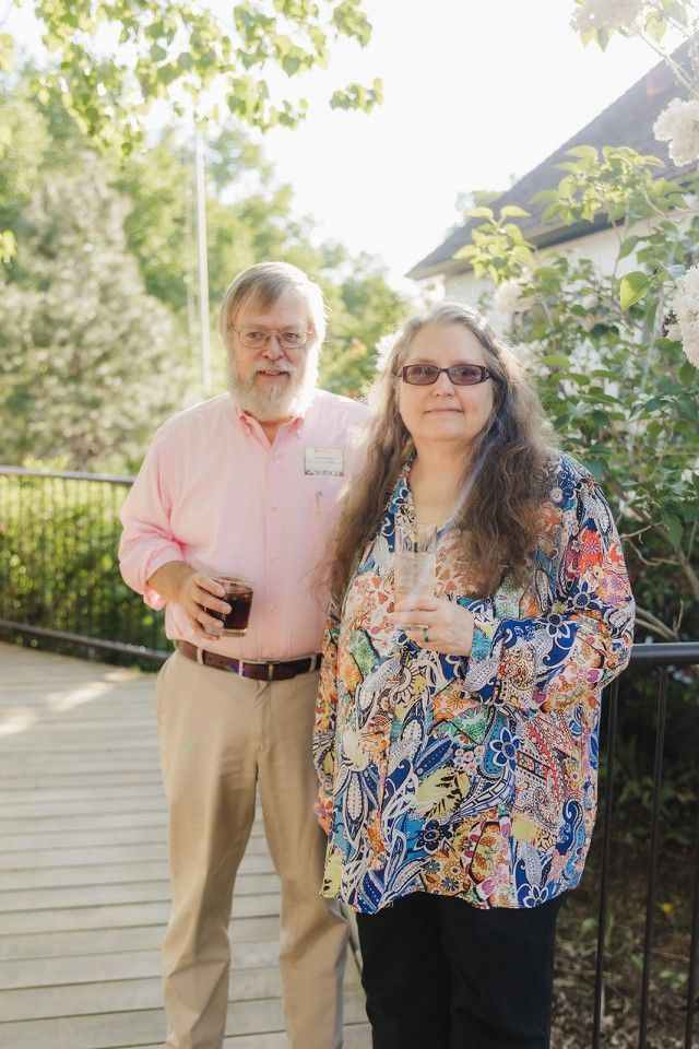 Couple standing outdoors on a wooden walkway. Man in pink shirt, woman in patterned top, holding drinks. Sunny day.