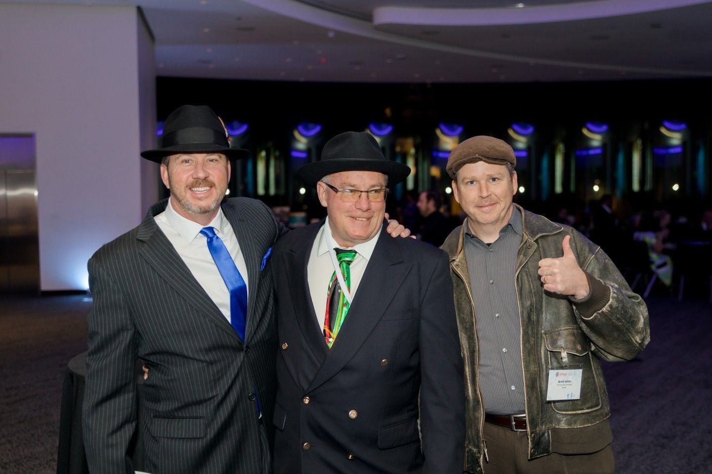 Three men in suits and hats pose indoors, smiling. One gives a thumbs-up.