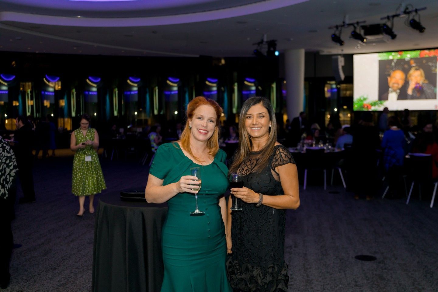 Two women at an event, one in green dress, the other in black, holding drinks. Event setting with lights.