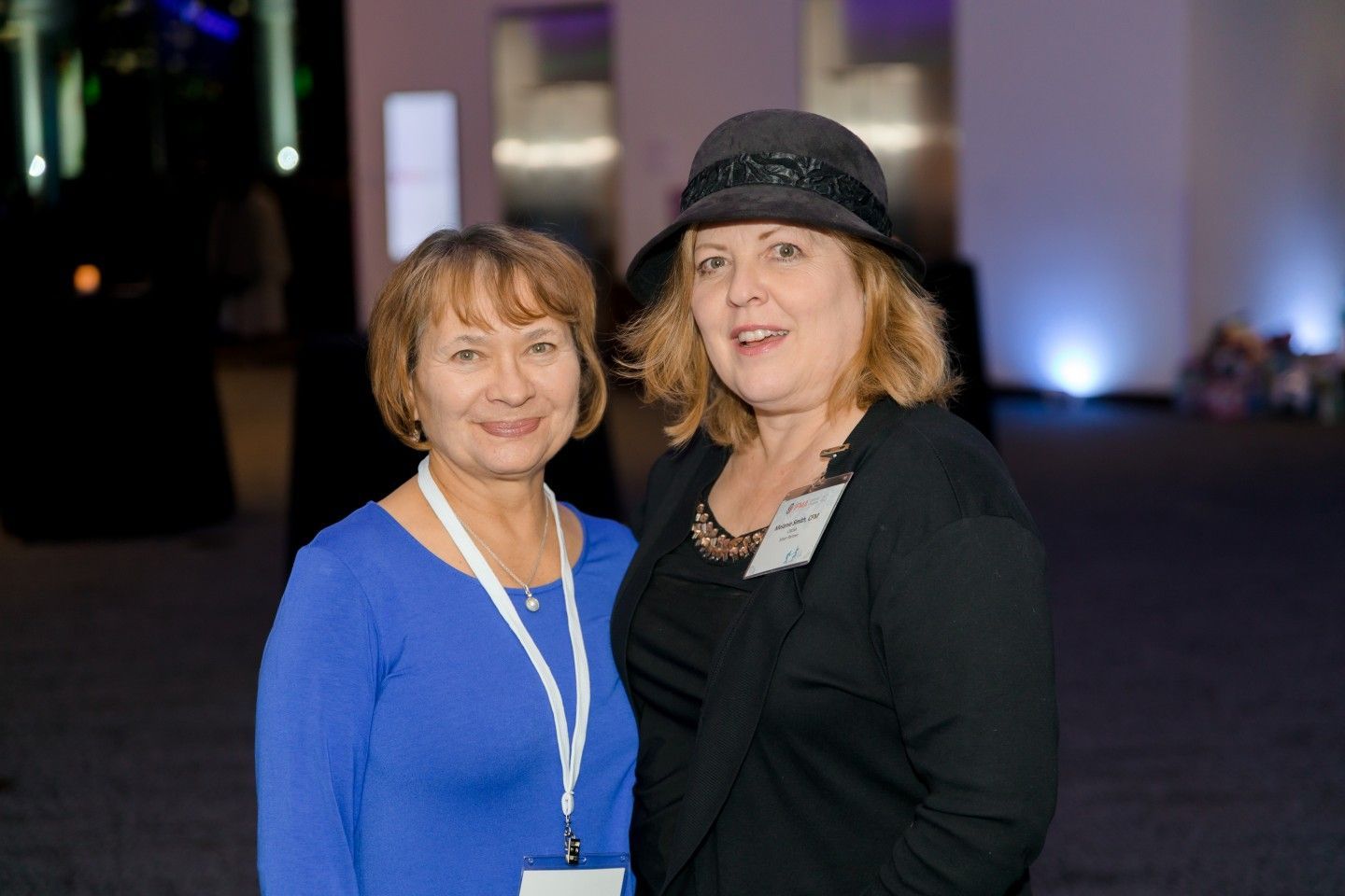 Two women smiling; one in blue shirt and white lanyard, the other in black, wearing a hat, posing together.