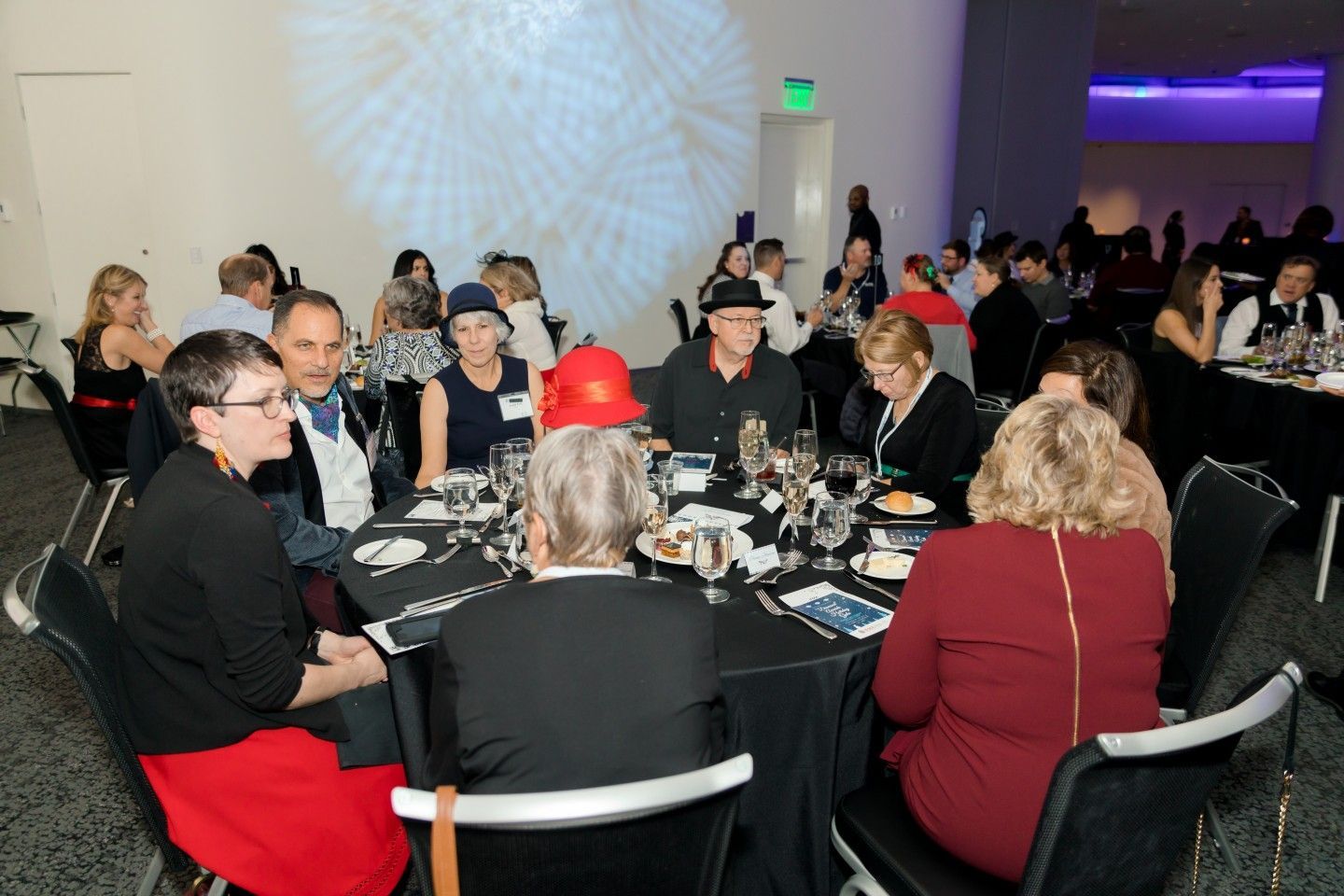 People seated at tables in a banquet hall, some wearing hats, under a projected light display.