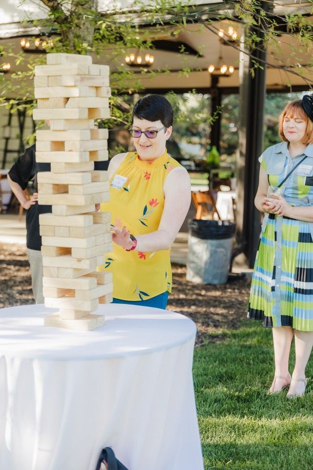 Woman in yellow shirt playing giant Jenga outdoors. Another person stands nearby.
