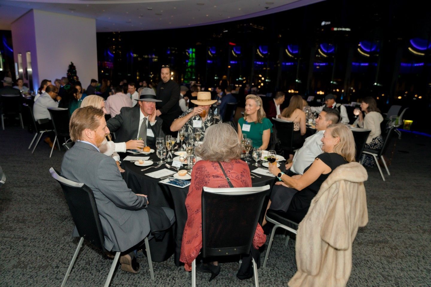 People seated at round tables in a dimly lit event space, with city lights visible outside.