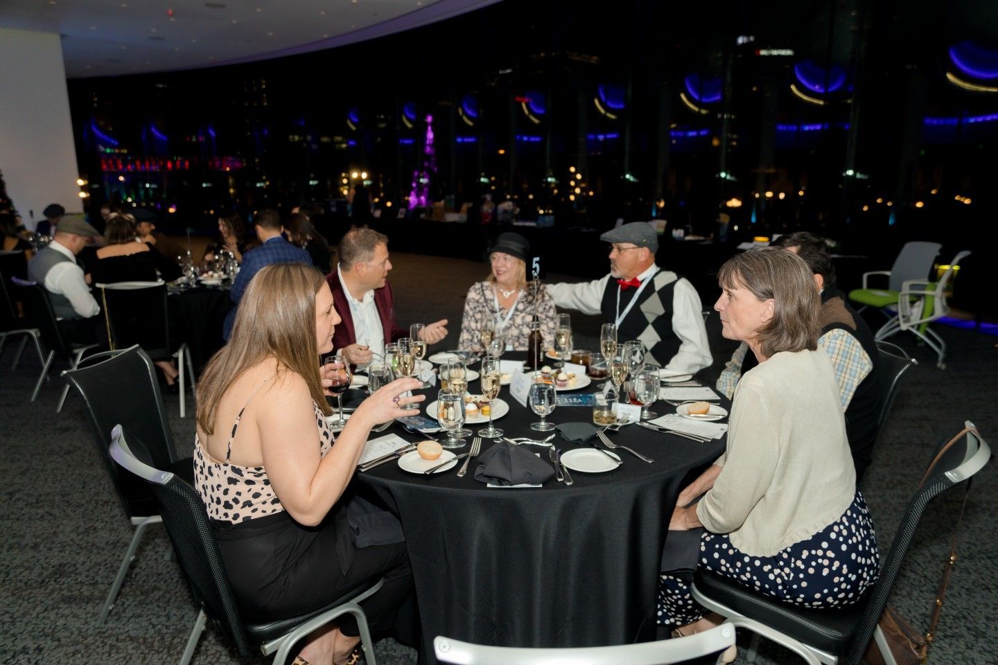 People seated at a round table, eating and conversing at a formal event, lit by ambient lighting.