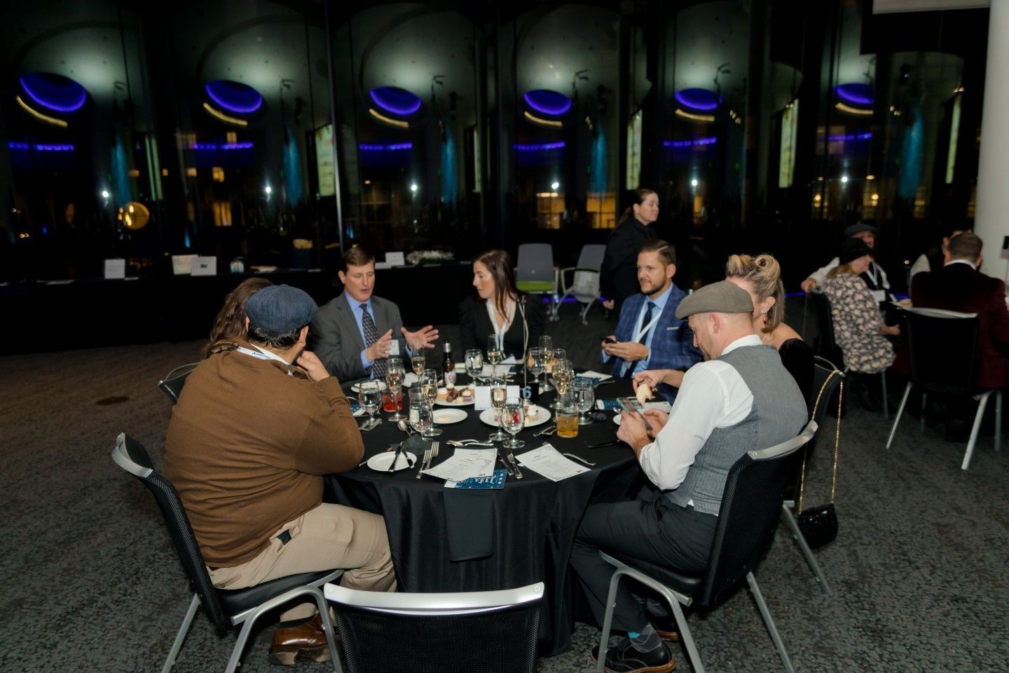 People seated at a round table, talking, in a dark room with background lights.