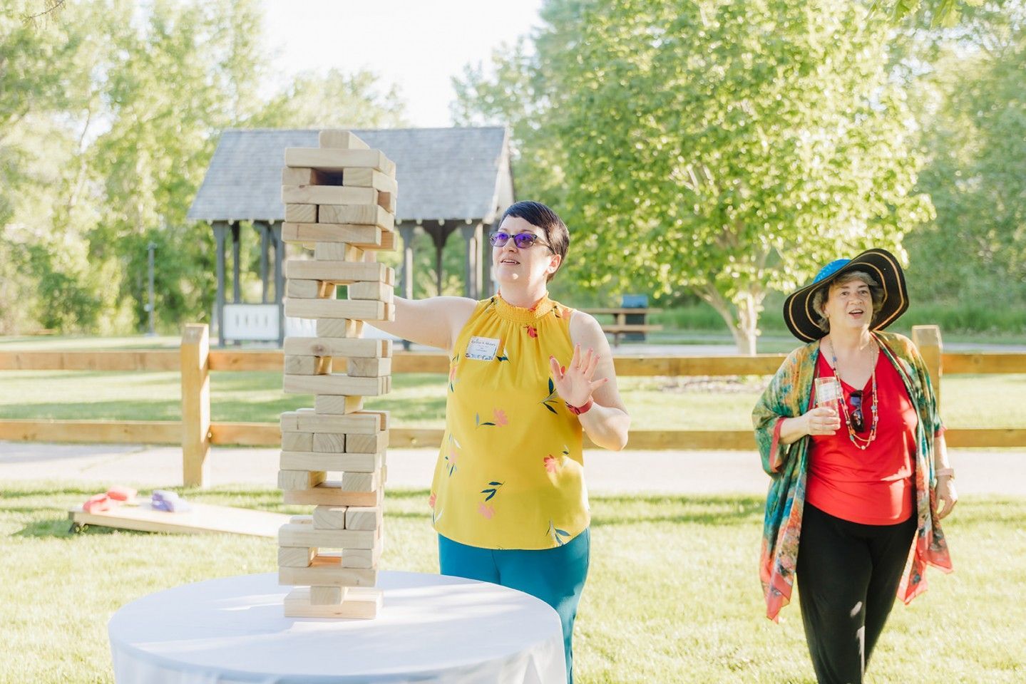 Two women play giant Jenga outdoors. One gestures towards the tower, the other watches.