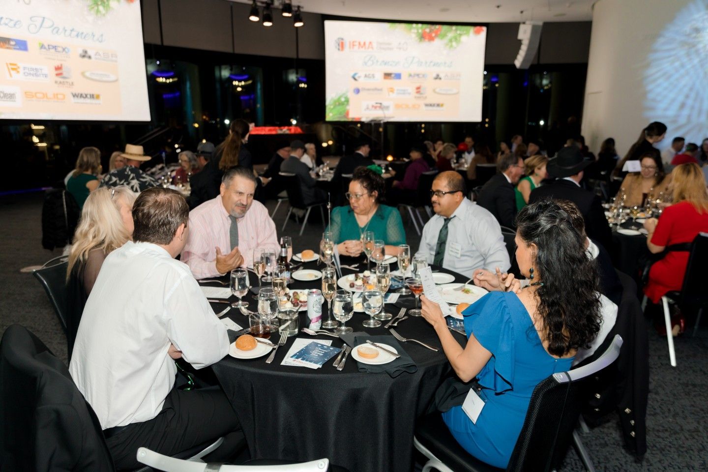 People seated at tables during a formal event, with two screens displaying logos and festive decor.