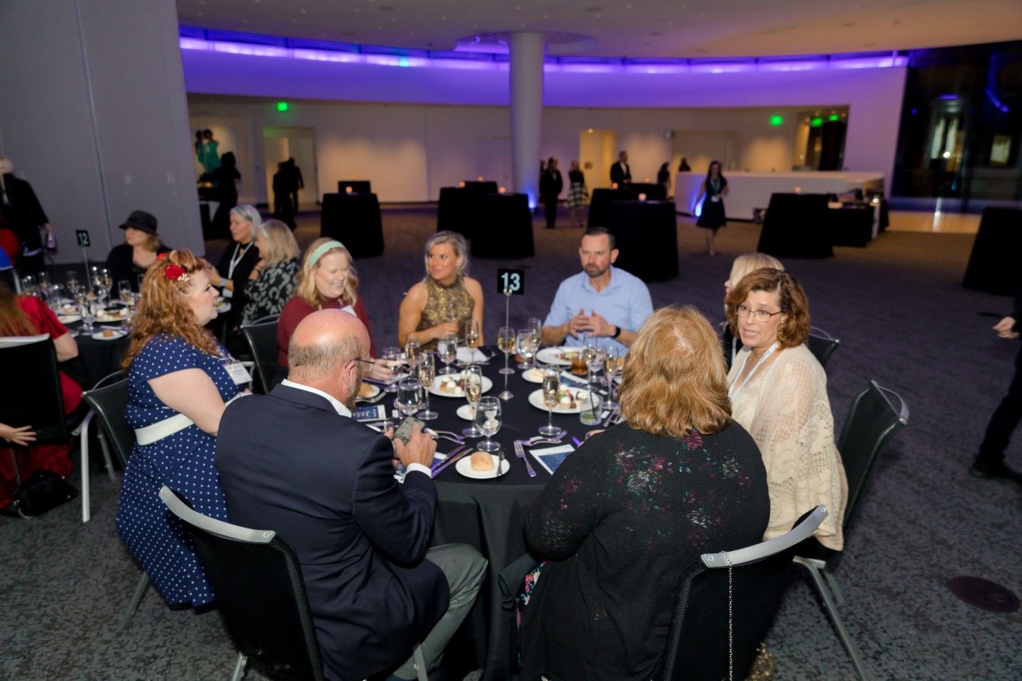 People seated at a round table, eating and conversing, in a large room with blue and purple lighting.