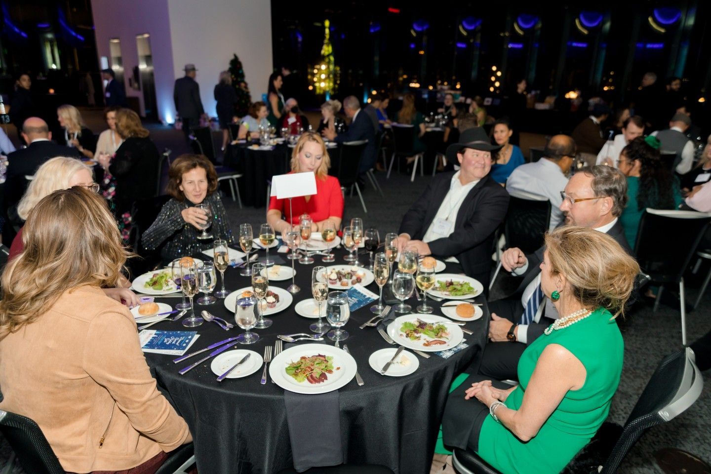 People seated at a round table at a formal event, eating and conversing. Dark room with other tables in background.