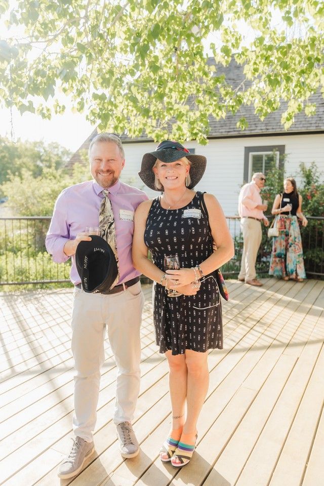 Man and woman pose on a deck. The woman wears a hat and dress, the man wears a tie and light pants.