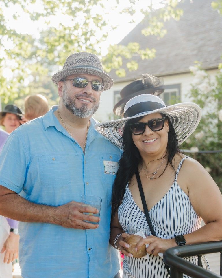 Couple wearing hats and sunglasses, smiling outdoors. Man in blue shirt, woman in striped dress.