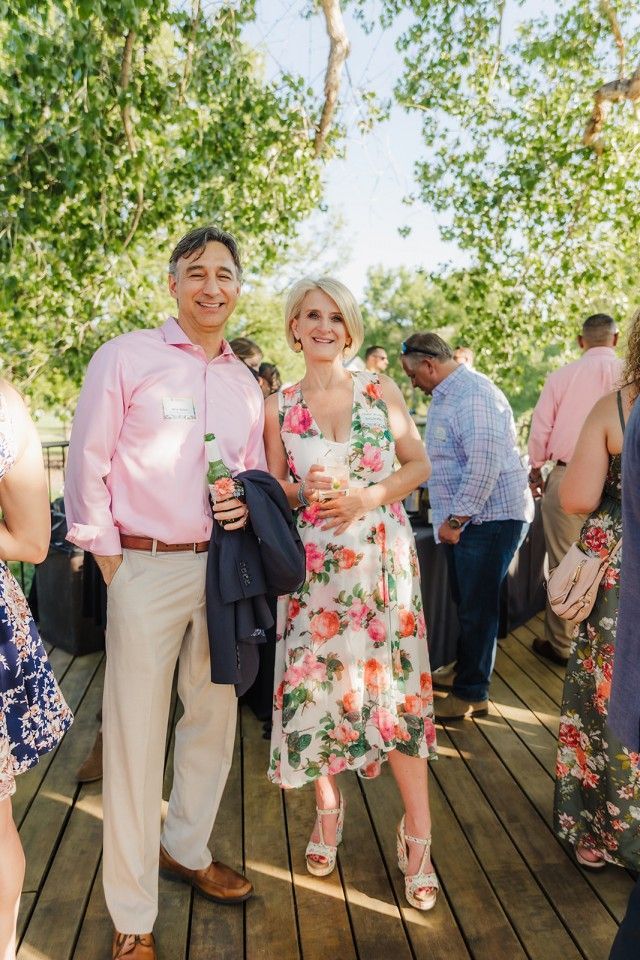 Man and woman at an outdoor event. Man in pink shirt, woman in floral dress, standing on a wooden deck.