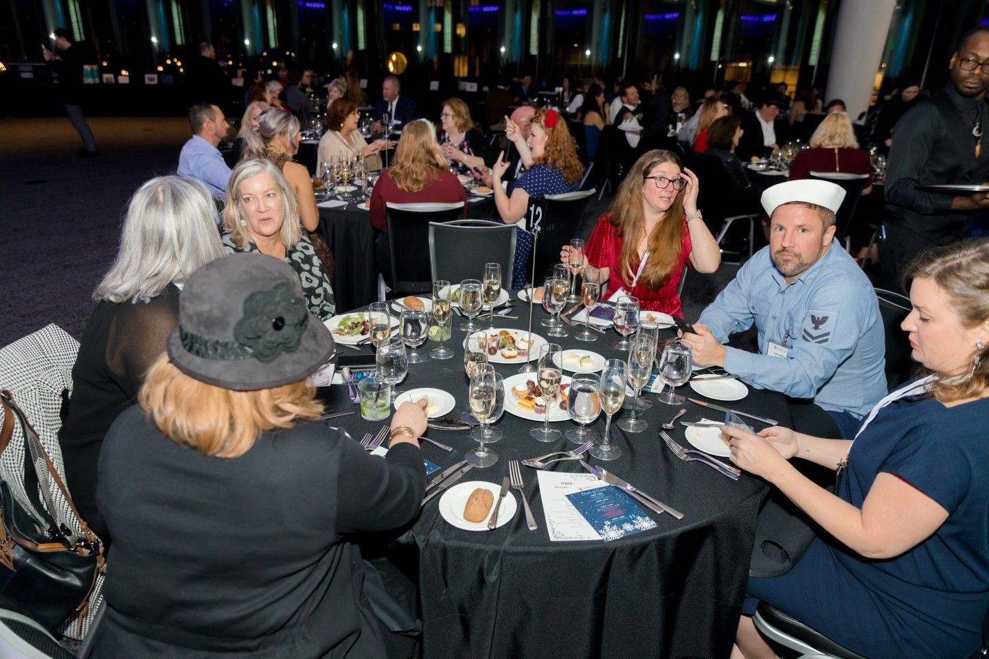 People seated around tables at a formal event, eating and talking. Dark tablecloths.