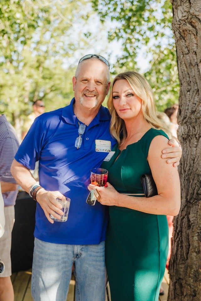 Man in blue shirt and woman in green dress embrace outdoors, both holding drinks.