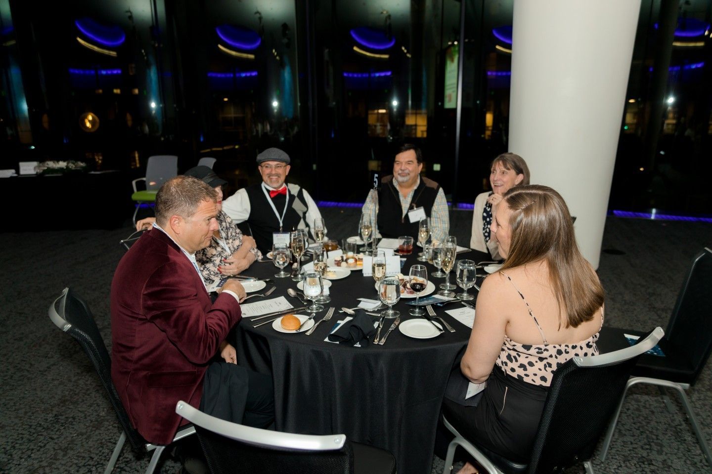 Six people seated at a round table, eating and conversing at a formal event, night setting.