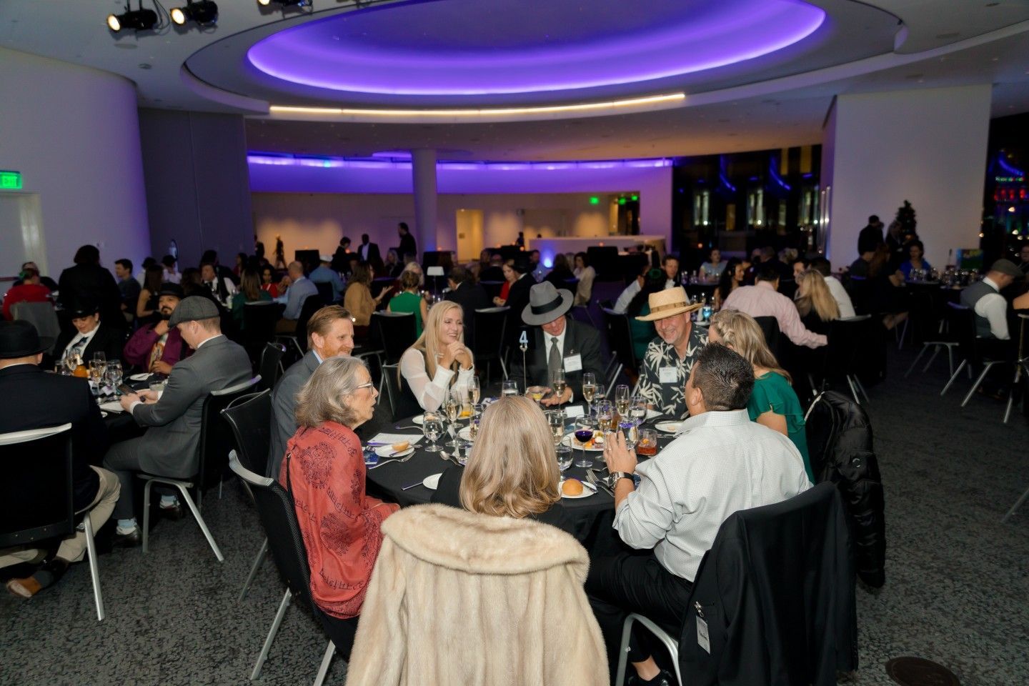 People seated at tables in a brightly lit event space with a purple-lit ceiling.