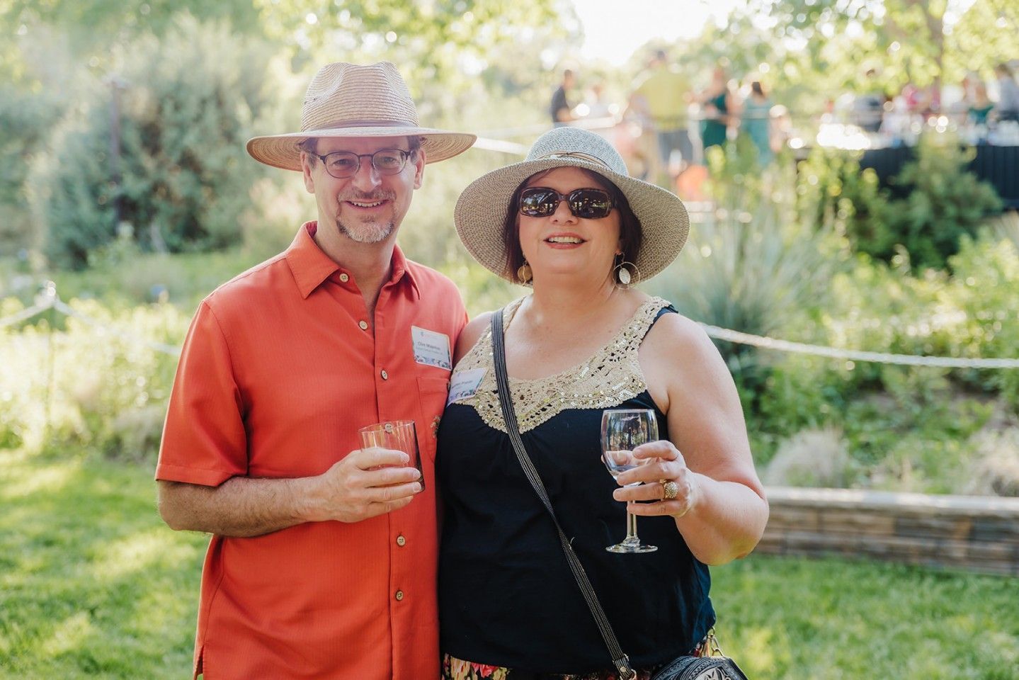 Couple smiling, holding drinks, in a garden. Man in orange shirt and straw hat. Woman in black top and hat.