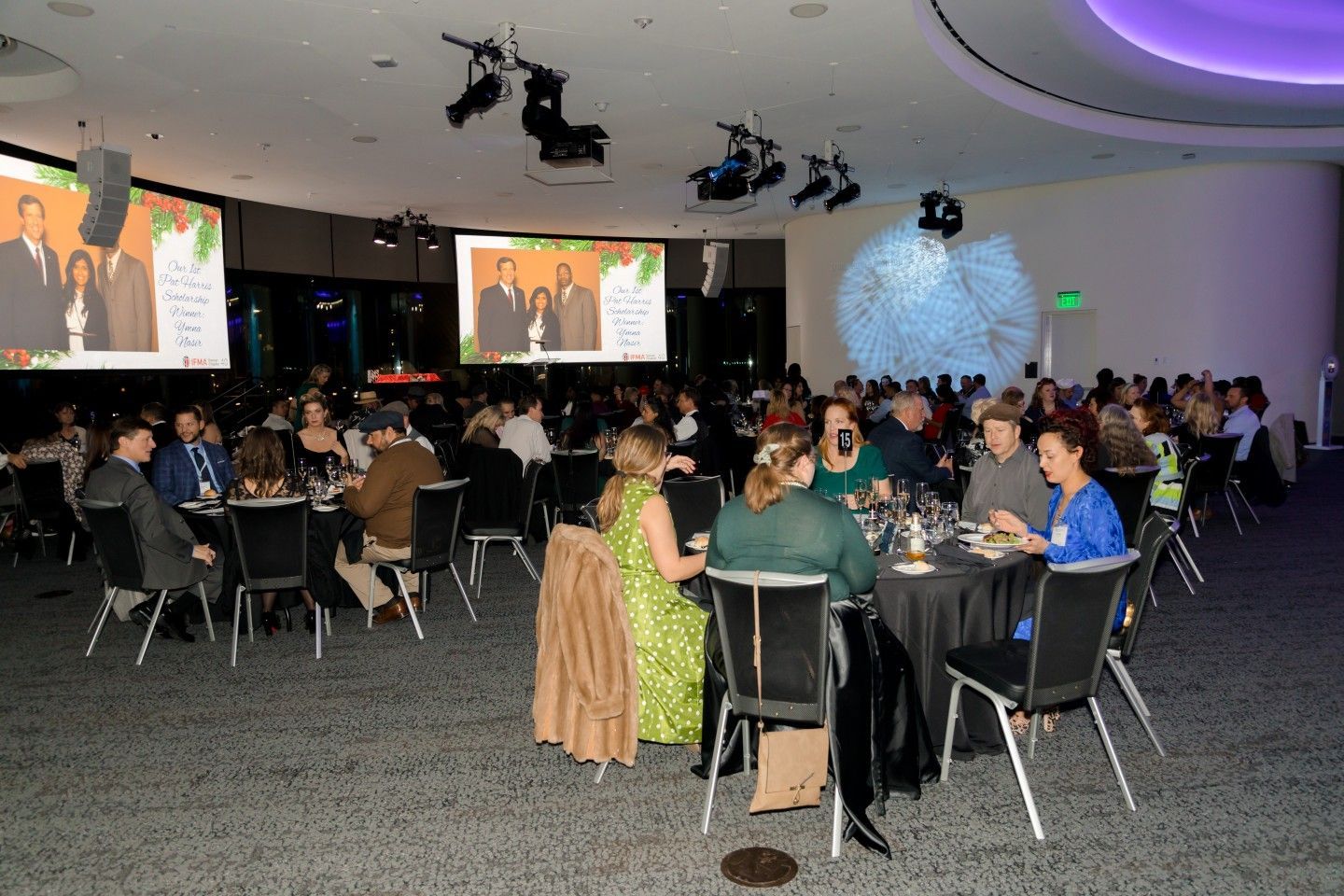 People seated at round tables in a large event space, watching screens.