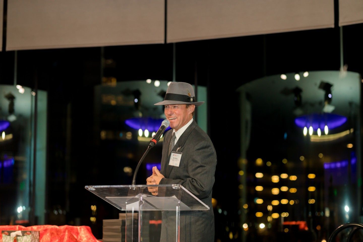 Man in suit and fedora speaks at a podium, night cityscape in the background.