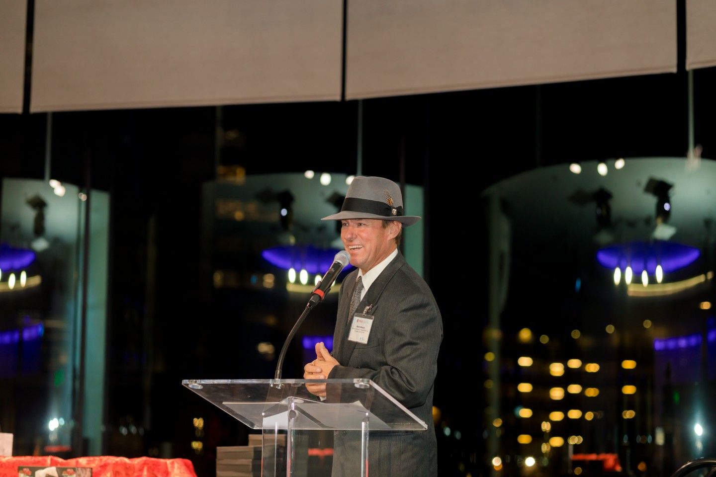 Man in hat speaks at podium, gesturing. Nighttime city background.