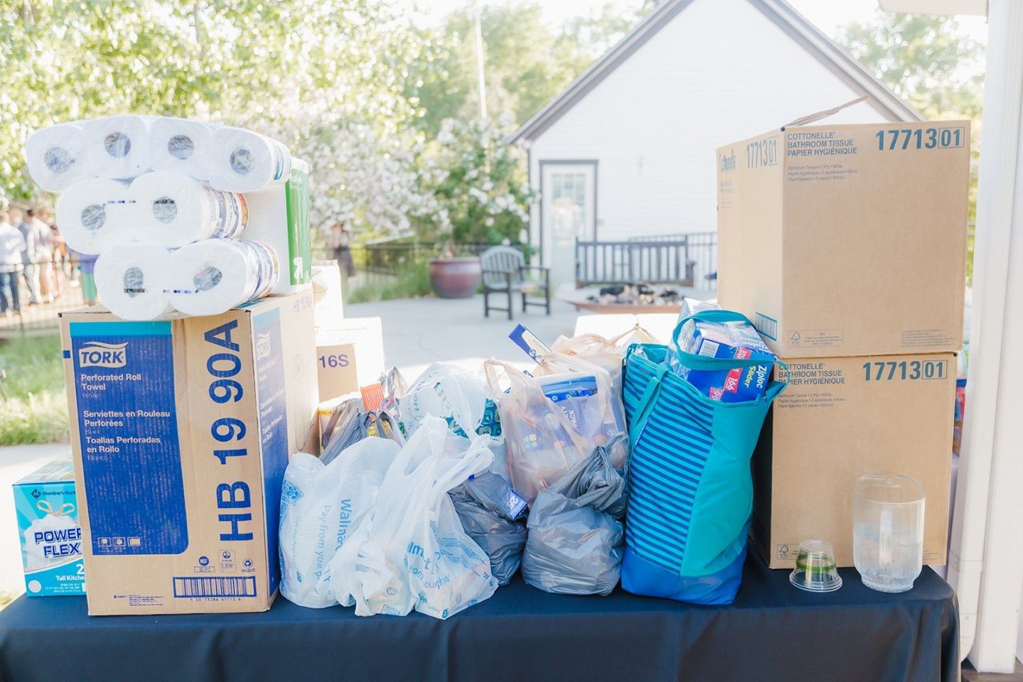 Table with donated supplies: toilet paper, paper towels, boxes, and plastic bags in front of a white building.