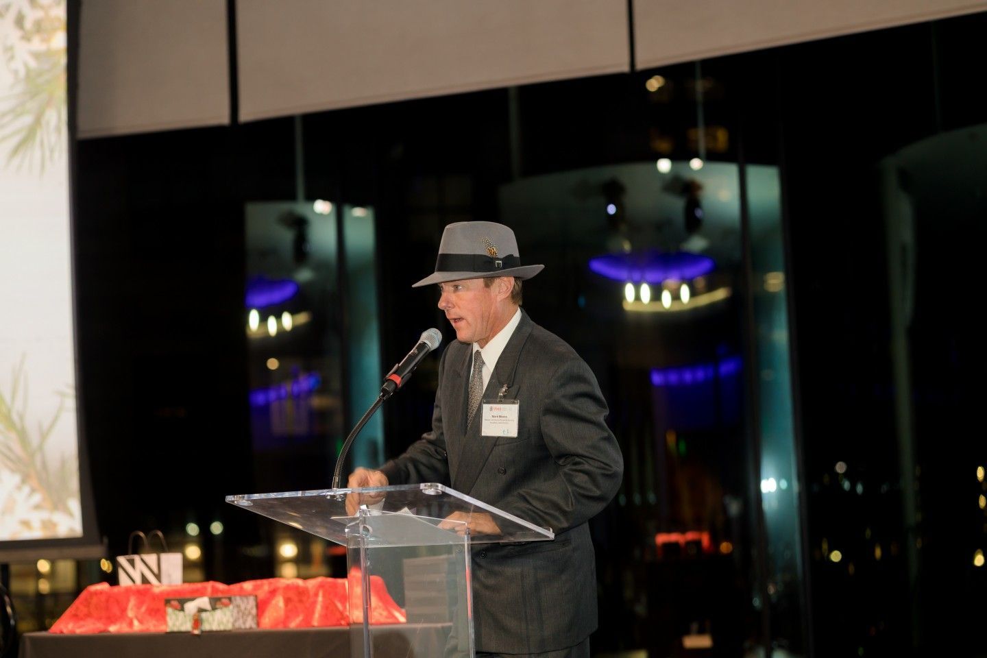 Man in suit and fedora speaks at a podium; city lights in background.