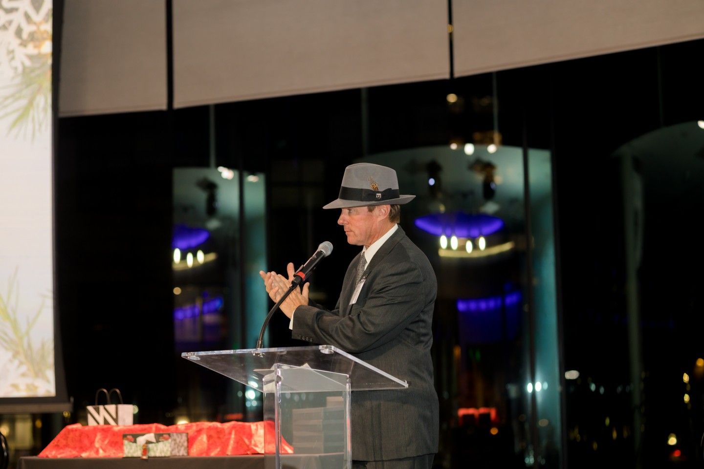Man in suit and fedora speaks at podium, clapping, in an event setting with city lights visible.