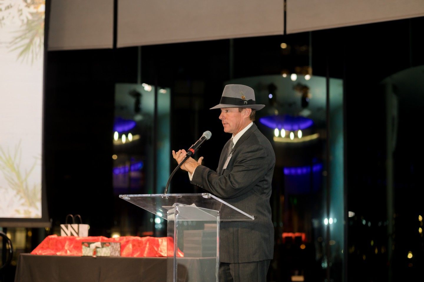 Man in suit and fedora speaks at podium, gesturing. Dark room, stage lighting.