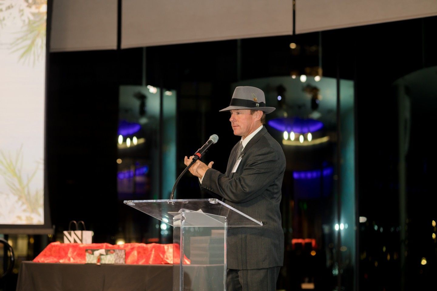 Man in suit and fedora speaks at podium, holding microphone, at night event.