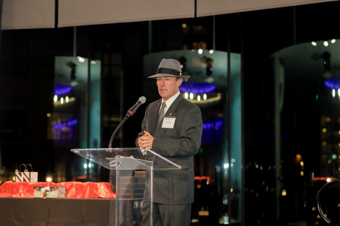 Man in suit and fedora speaks at podium with city lights in background.