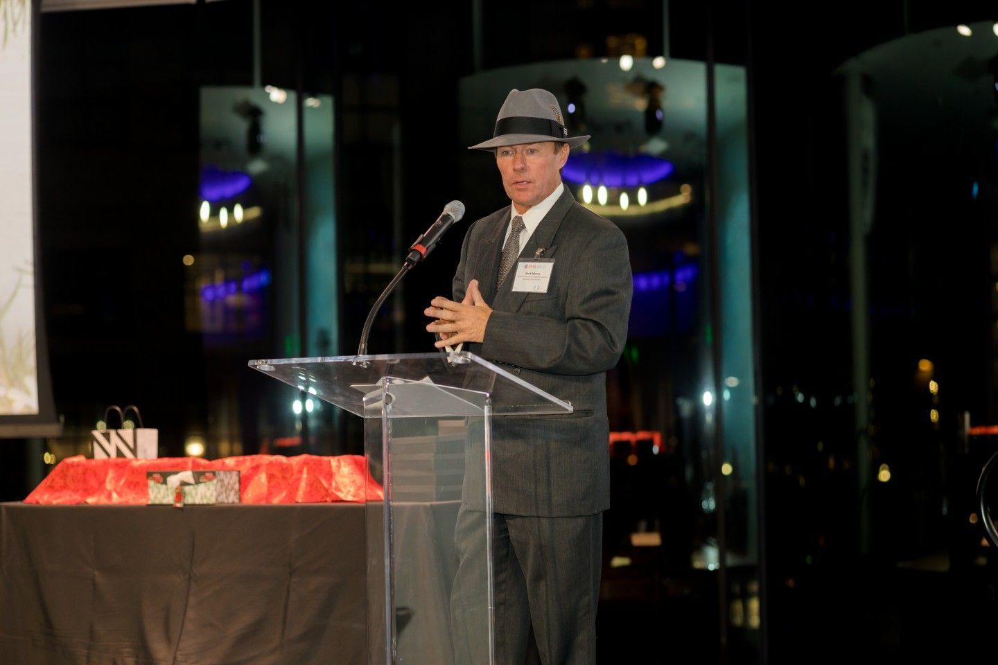 Man in fedora speaking at a podium with city lights in the background.