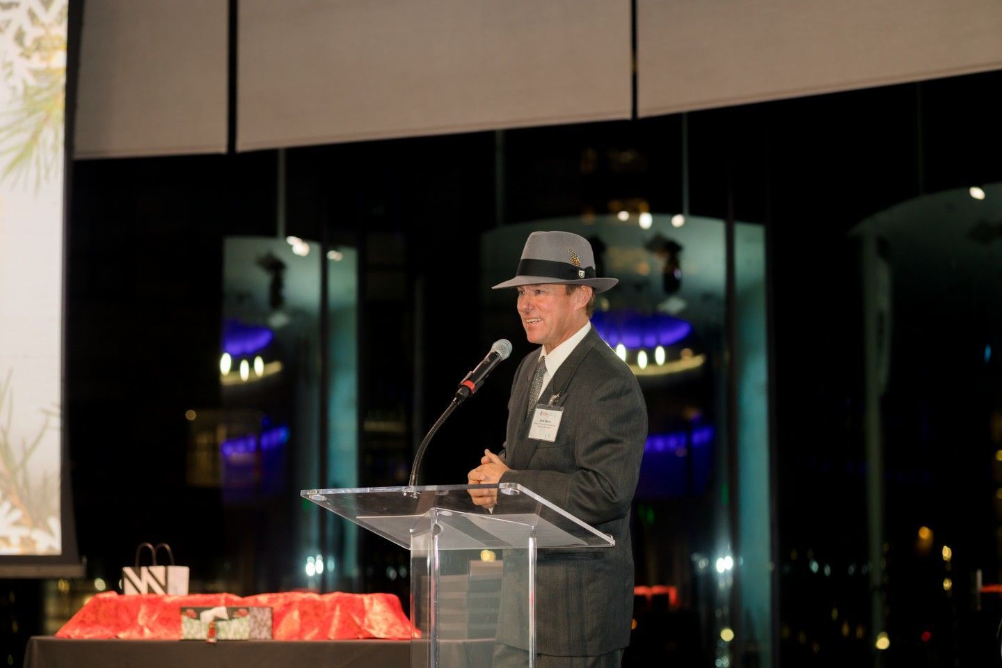 Man in a fedora speaks at a podium, night cityscape in the background.