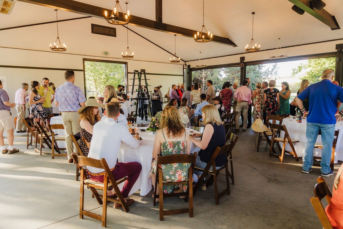 People at tables in an outdoor event space, some standing, some seated, with a neutral-colored ceiling and open sides.
