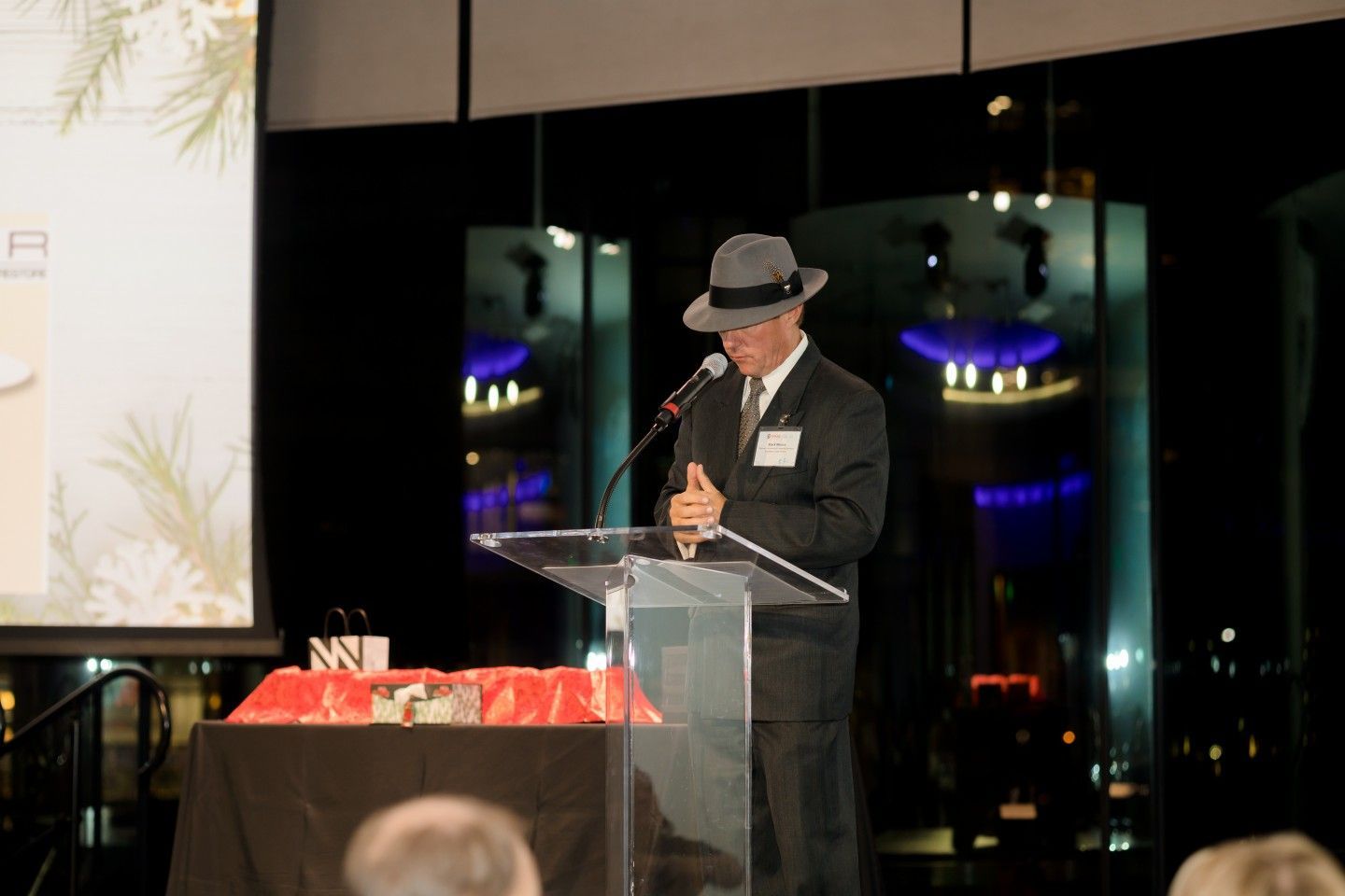 Man in suit and fedora speaks at a podium; formal event setting with decorations and screen.