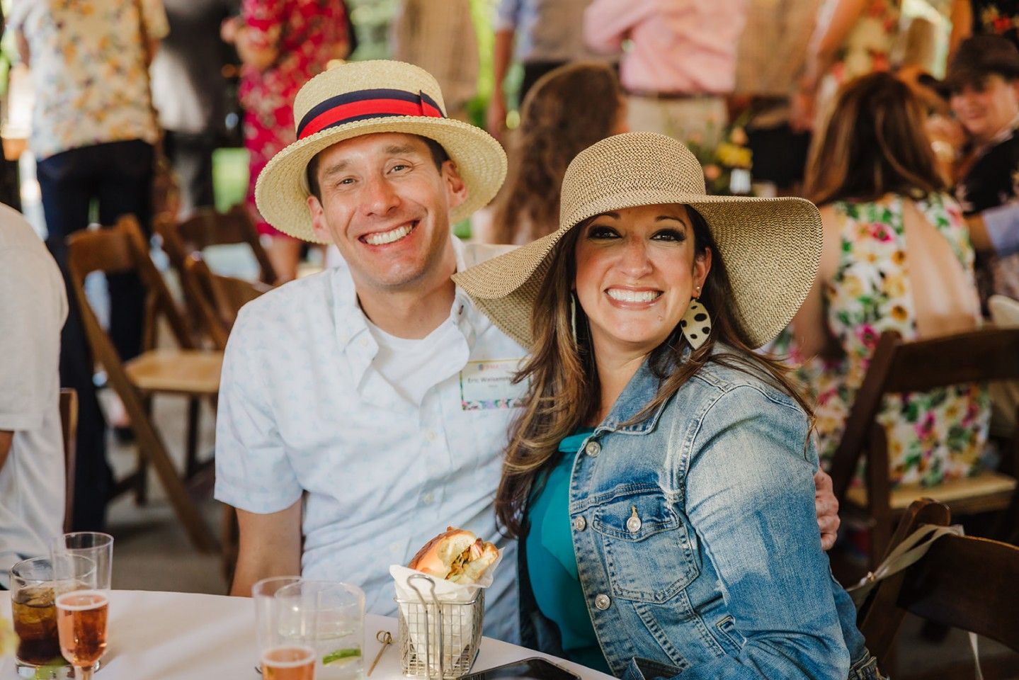 Couple wearing hats smiling, sitting at a table outdoors. Woman in denim jacket, man in white shirt, crowd in background.