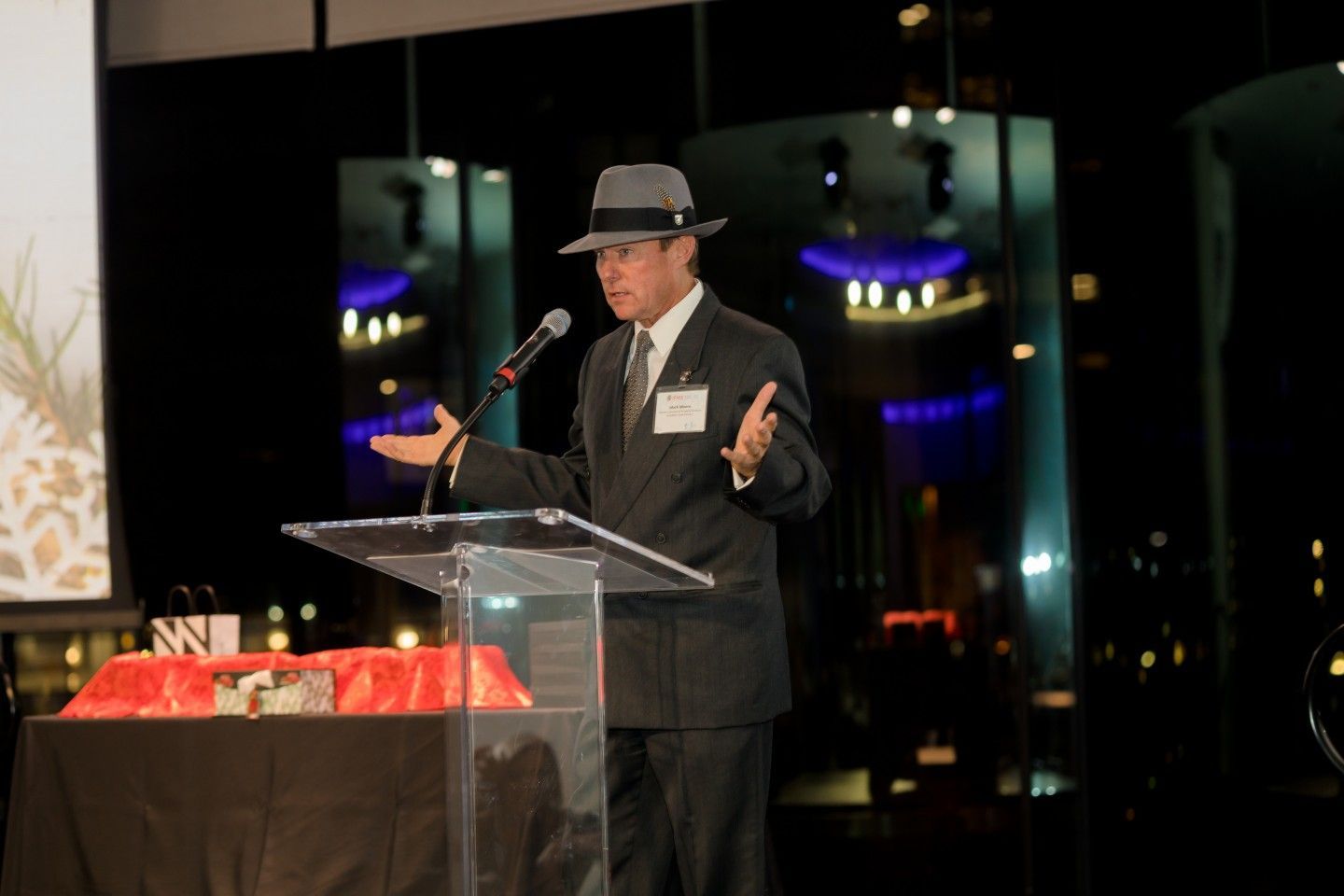 Man in suit and fedora speaks at podium during an evening event, arms raised, city lights in background.
