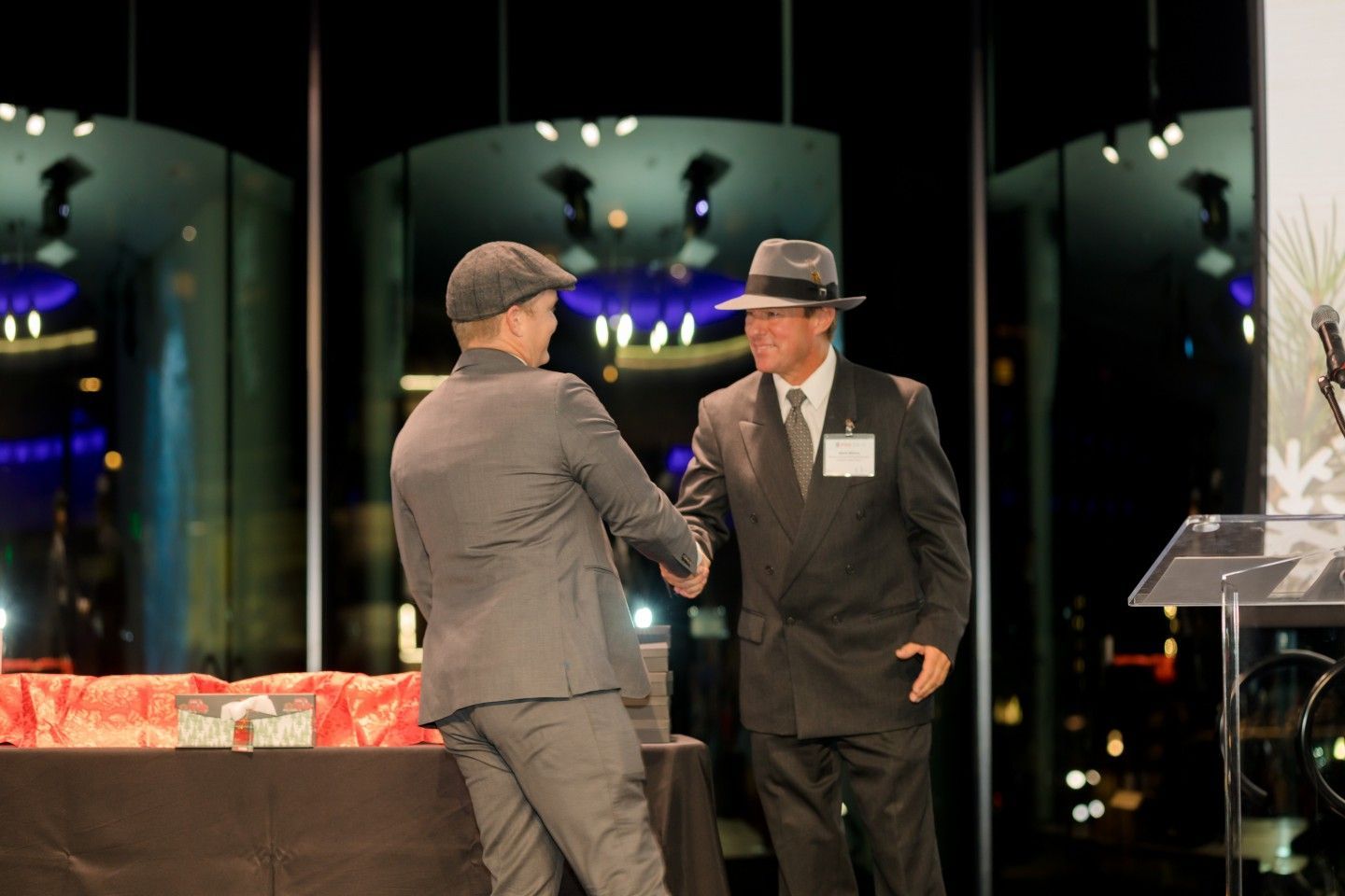 Two men shaking hands at an awards ceremony, both wearing hats and suits; night setting.