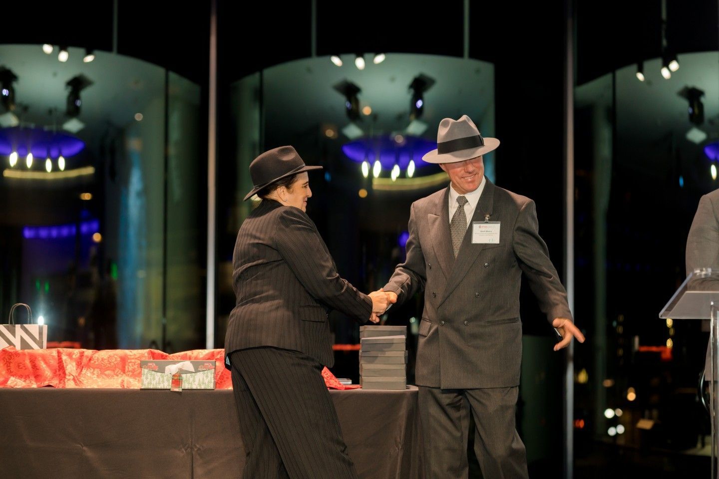 Two people in suits and fedoras shaking hands on a stage; a celebratory event.