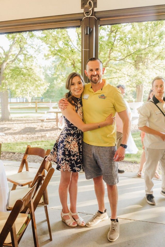 Woman in floral dress hugs man in yellow shirt and shorts; outdoor event setting.