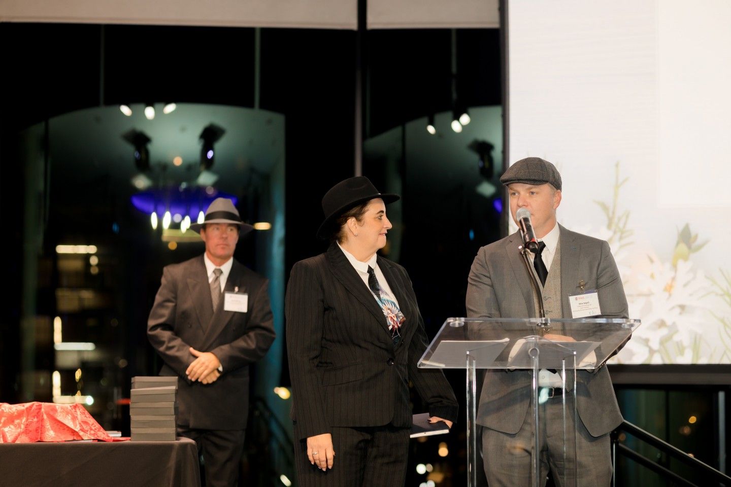 Three people in vintage attire on stage, one speaking at a podium. Nighttime background with city lights.