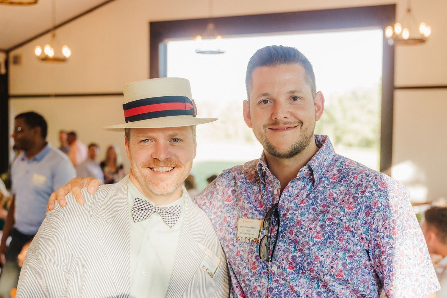 Two men smiling, one in a seersucker suit and straw hat, the other in a floral shirt. Indoors.