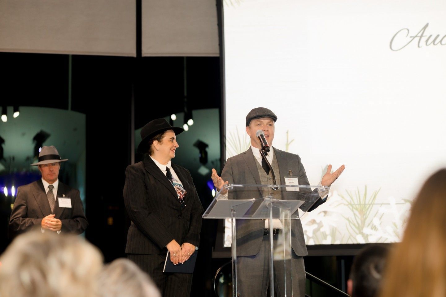 People at podium with microphone; speaking at event; woman in suit, man in suit and hat.