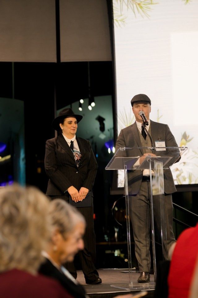 Two people in suits at a podium, one speaking into a microphone, the other standing beside him; indoors, audience visible.